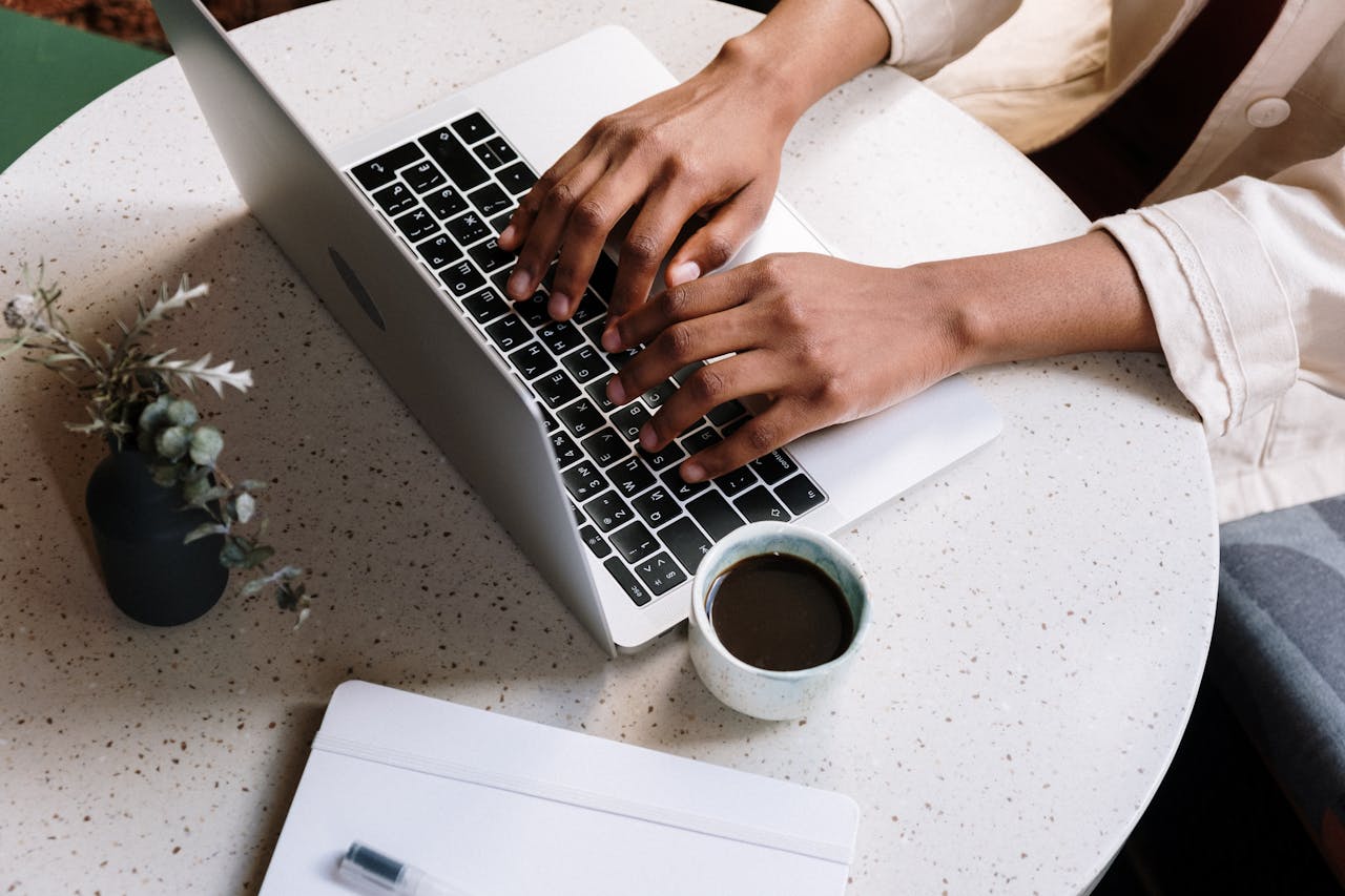 discover A person typing on a laptop at a café table with coffee and a notebook.